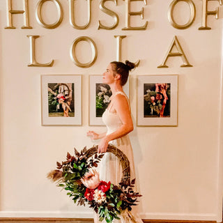 Woman holding a floral wreath in front of 'HOUSE OF LO LA' sign